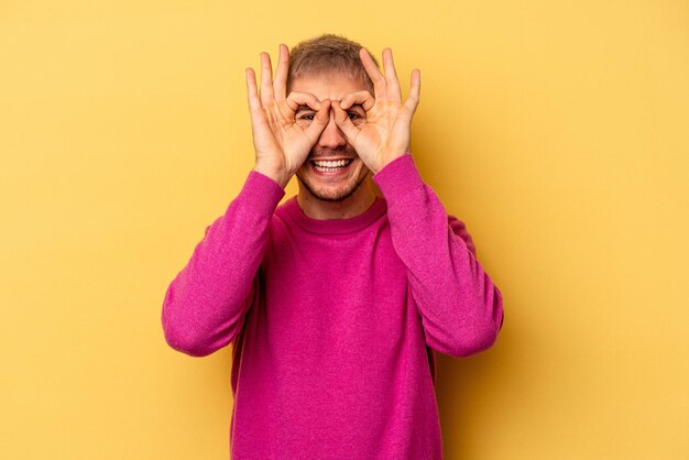 Hombre caucásico joven aislado sobre fondo amarillo que muestra un buen signo sobre los ojos