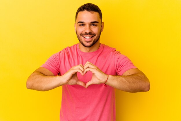 Hombre caucásico joven aislado sobre fondo amarillo sonriendo y mostrando una forma de corazón con las manos.