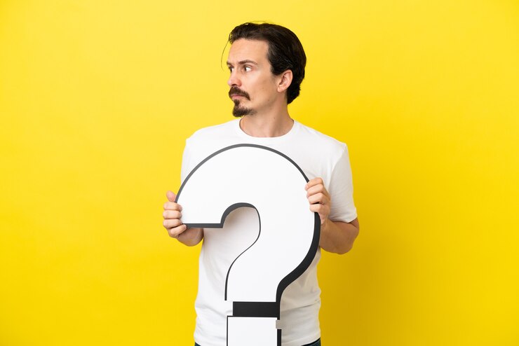 Foto hombre caucásico joven aislado sobre fondo amarillo sosteniendo un icono de signo de interrogación y mirando de lado