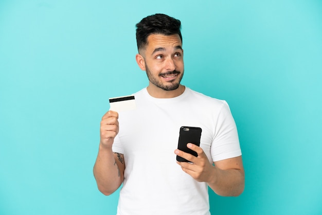 Hombre caucásico joven aislado sobre fondo azul comprando con el móvil con una tarjeta de crédito mientras piensa