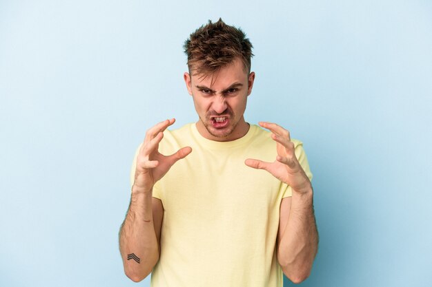 Foto hombre caucásico joven aislado sobre fondo azul molesto gritando con las manos tensas.