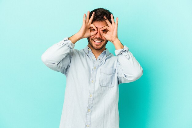 Hombre caucásico joven aislado sobre fondo azul que muestra bien firmar sobre los ojos