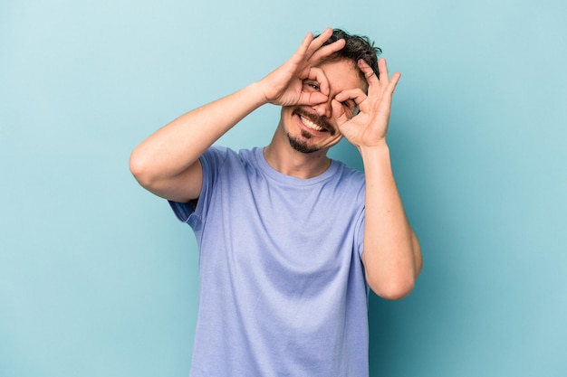 Hombre caucásico joven aislado sobre fondo azul que muestra un buen signo sobre los ojos