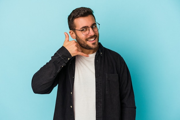 Hombre caucásico joven aislado sobre fondo azul que muestra un gesto de llamada de teléfono móvil con los dedos.