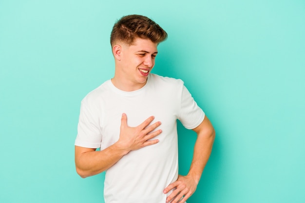 Hombre caucásico joven aislado sobre fondo azul riendo manteniendo las manos en el corazón, concepto de felicidad.