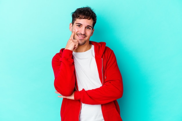 Hombre caucásico joven aislado sobre fondo azul sonriendo feliz y confiado, tocando la barbilla con la mano.