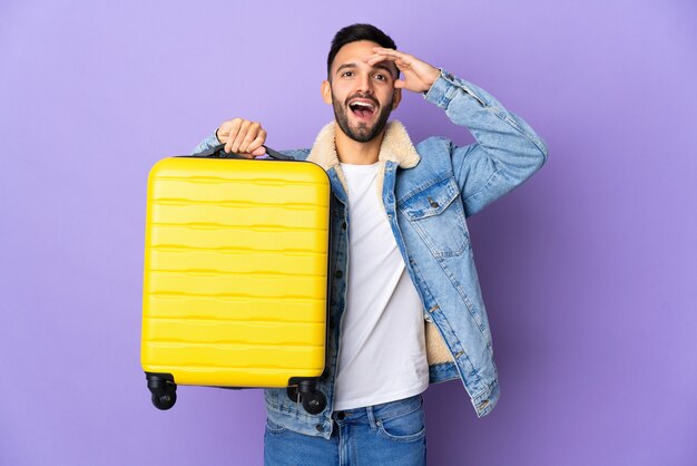 Hombre caucásico joven aislado sobre fondo azul en vacaciones con maleta de viaje y sorprendido