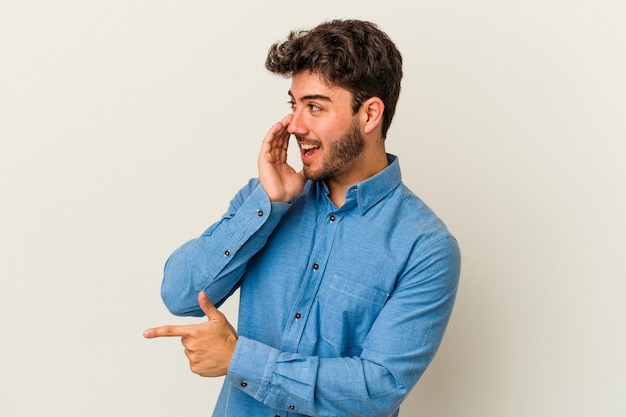 Foto hombre caucásico joven aislado sobre fondo blanco diciendo un chisme, apuntando al lado informando algo.