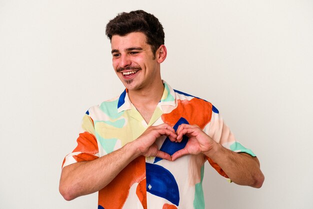 Hombre caucásico joven aislado sobre fondo blanco sonriendo y mostrando una forma de corazón con las manos.