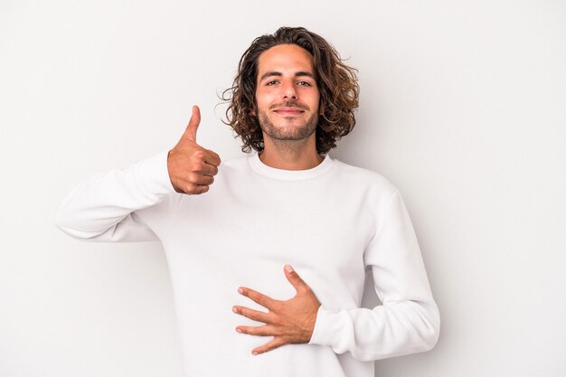 Hombre caucásico joven aislado sobre fondo gris toca la barriga, sonríe suavemente, comiendo y concepto de satisfacción.