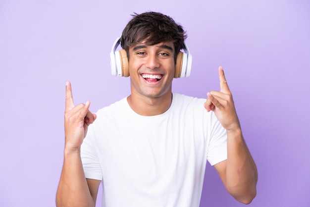 Hombre caucásico joven aislado sobre fondo púrpura escuchando música haciendo gesto de rock