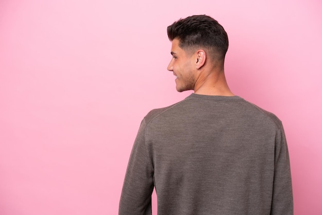 Hombre caucásico joven aislado sobre fondo rosa en posición de espalda y mirando de lado