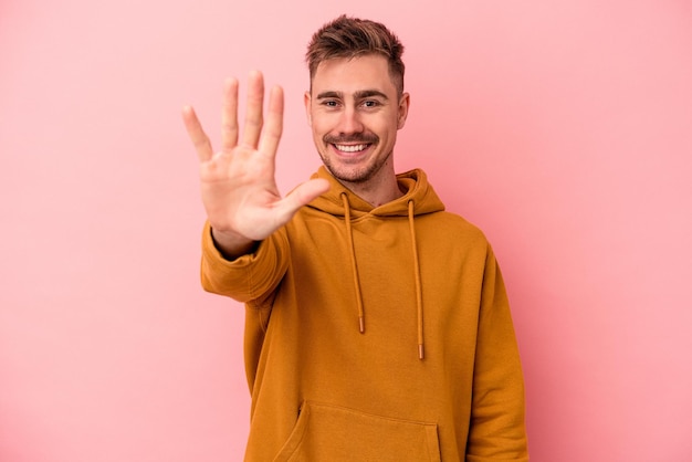 Hombre caucásico joven aislado sobre fondo rosa sonriendo alegre mostrando el número cinco con los dedos.