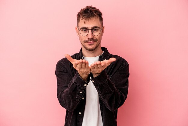 Hombre caucásico joven aislado sobre fondo rosa sosteniendo algo con las palmas, ofreciendo a la cámara.