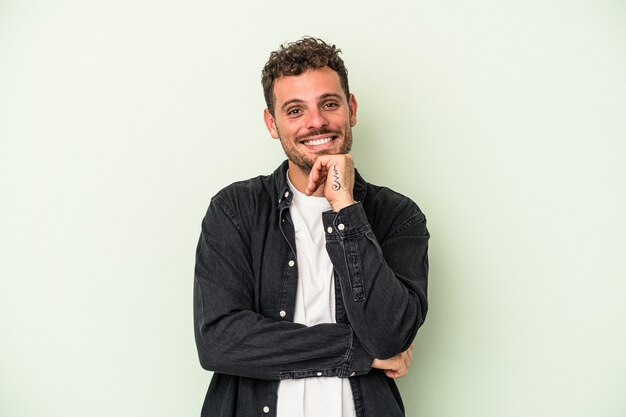 Hombre caucásico joven aislado sobre fondo verde sonriendo feliz y confiado, tocando la barbilla con la mano.