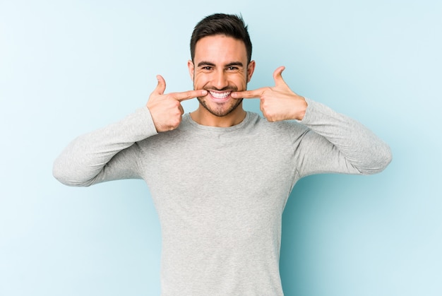 Foto hombre caucásico joven aislado en sonrisas azules, señalando con el dedo en la boca.