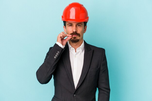 Hombre caucásico joven arquitecto aislado sobre fondo azul con los dedos en los labios manteniendo un secreto.