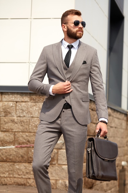 Hombre caucásico joven con barba en traje junto al edificio después de la reunión de negocios, chico en esmoquin y gafas de sol negras al aire libre. concepto de gente de negocios