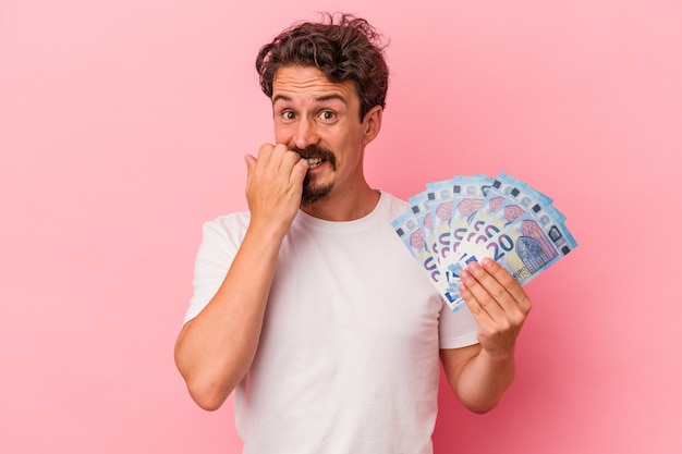 Hombre caucásico joven con billetes aislados sobre fondo rosa mordiéndose las uñas, nervioso y muy ansioso.