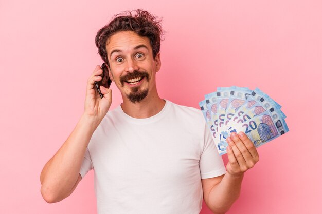 Hombre caucásico joven con billetes y hucha aislado sobre fondo rosa