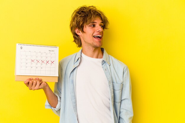 Hombre caucásico joven con calendario de celebración de maquillaje aislado sobre fondo amarillo se ve a un lado sonriente, alegre y agradable.