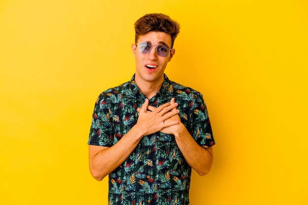 Hombre caucásico joven con una camisa hawaiana aislada sobre fondo amarillo tiene una expresión amistosa, presionando la palma contra el pecho. Concepto de amor.