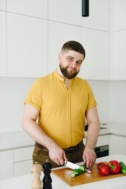 Foto hombre caucásico joven en camiseta amarilla que cocina la ensalada o la comida vegetal sana en la cocina moderna blanca con un cuchillo que sonríe