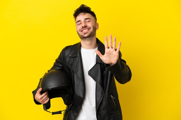 Hombre caucásico joven con un casco de motocicleta aislado sobre fondo amarillo contando cinco con los dedos