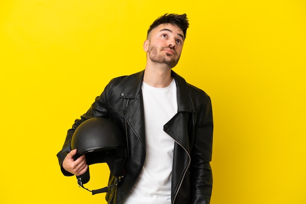 Hombre caucásico joven con un casco de motocicleta aislado sobre fondo amarillo y mirando hacia arriba