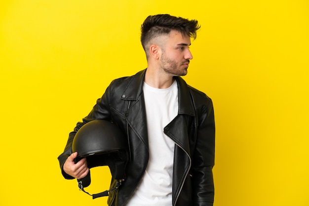 Hombre caucásico joven con un casco de motocicleta aislado sobre fondo amarillo mirando hacia el lado