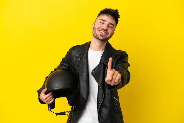 Hombre caucásico joven con un casco de motocicleta aislado sobre fondo amarillo mostrando y levantando un dedo