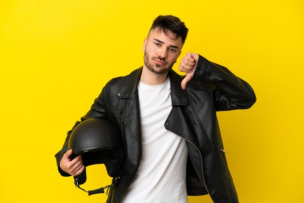 Hombre caucásico joven con un casco de motocicleta aislado sobre fondo amarillo que muestra el pulgar hacia abajo con expresión negativa