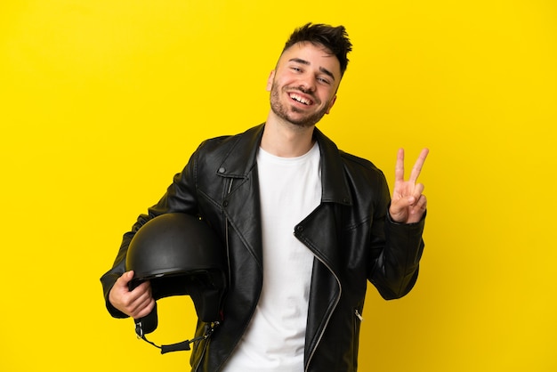 Hombre caucásico joven con un casco de motocicleta aislado sobre fondo amarillo sonriendo y mostrando el signo de la victoria