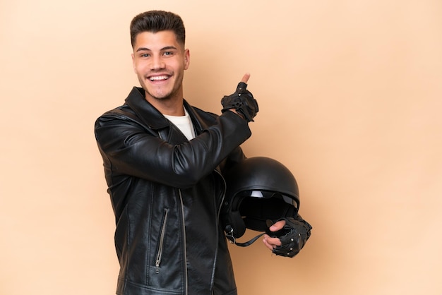 Hombre caucásico joven con un casco de motocicleta aislado sobre fondo beige apuntando hacia atrás