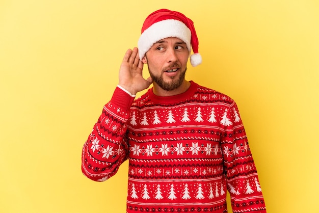 Hombre caucásico joven celebrando la Navidad aislado sobre fondo amarillo tratando de escuchar un chisme.