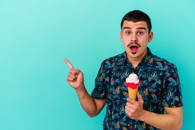 Hombre caucásico joven comiendo un helado aislado en azul apuntando hacia el lado