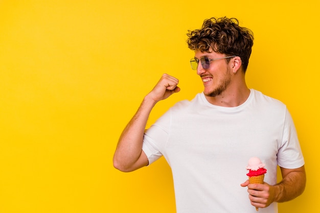 Hombre caucásico joven comiendo un helado aislado sobre fondo amarillo levantando el puño después de una victoria, concepto ganador.