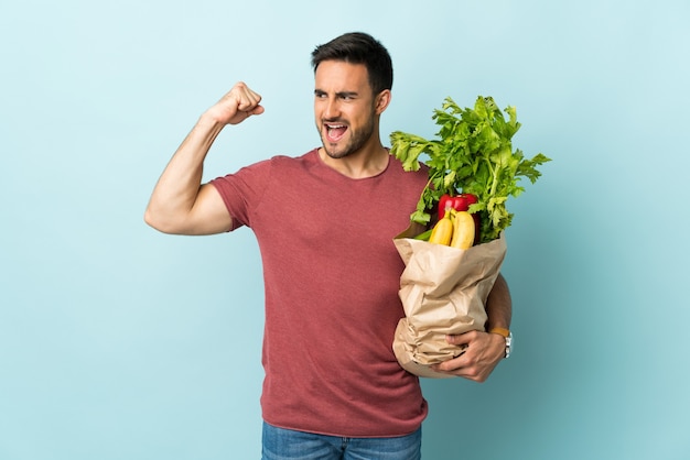 Hombre caucásico joven comprando algunas verduras aisladas en azul celebrando una victoria