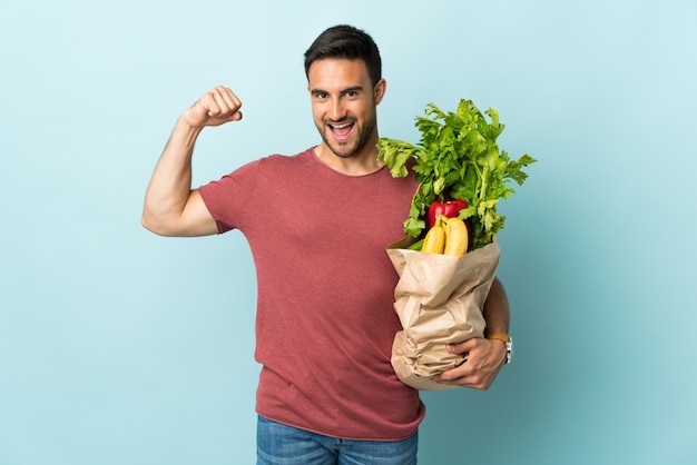 Hombre caucásico joven comprando algunas verduras aisladas en azul haciendo un gesto fuerte