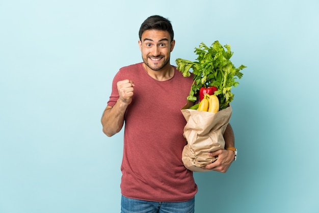 Hombre caucásico joven comprando algunas verduras aisladas sobre fondo azul celebrando una victoria