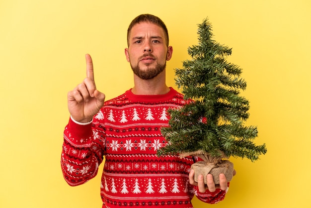 Hombre caucásico joven comprando arbolito para Navidad aislado sobre fondo amarillo mostrando el número uno con el dedo.