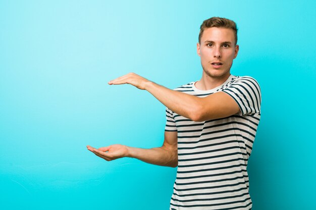 Hombre caucásico joven contra una pared azul conmocionado y sorprendido sosteniendo un espacio de copia entre las manos.