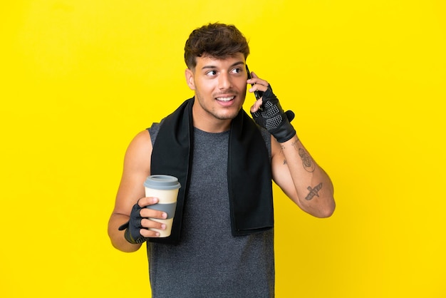 Hombre caucásico joven deporte con toalla aislado sobre fondo amarillo sosteniendo café para llevar y un móvil