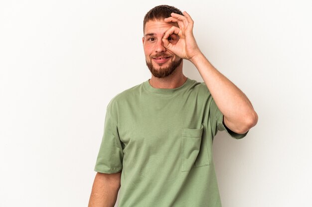 Foto hombre caucásico joven con diastema aislado sobre fondo blanco emocionado manteniendo el gesto ok en el ojo.