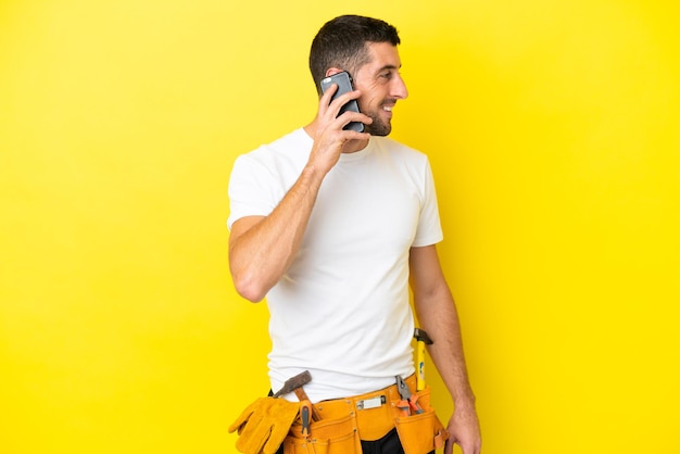 Hombre caucásico joven electricista aislado sobre fondo amarillo manteniendo una conversación con el teléfono móvil con alguien