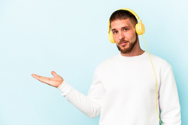 Hombre caucásico joven escuchando música aislada sobre fondo azul mostrando un espacio de copia en una palma y sosteniendo otra mano en la cintura.