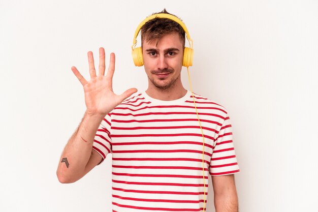 Hombre caucásico joven escuchando música aislada sobre fondo blanco sonriendo alegre mostrando el número cinco con los dedos.