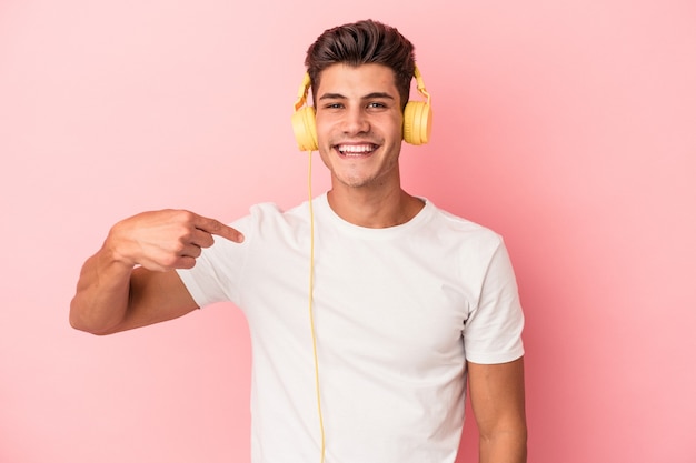 Hombre caucásico joven escuchando música aislada sobre fondo rosa persona apuntando con la mano a un espacio de copia de camisa, orgulloso y seguro