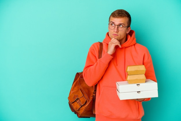 Hombre caucásico joven estudiante sosteniendo hamburguesas y pizzas aisladas en azul mirando hacia los lados con expresión dudosa y escéptica.