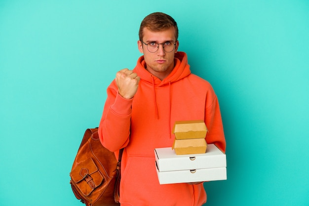 Hombre caucásico joven estudiante sosteniendo hamburguesas y pizzas aisladas en azul mostrando puño, expresión facial agresiva.